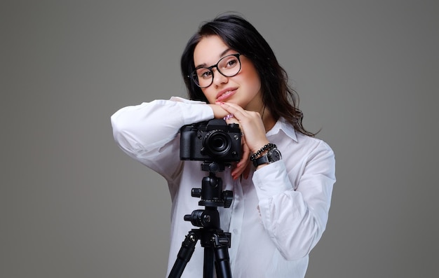 Smiling, positive brunette female in eyeglasses taking pictures with a professional photo camera. Isolated on grey background.