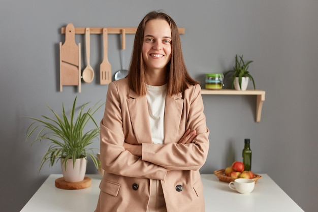 Улыбающаяся позитивная деловая женщина с каштановыми волосами в бежевой куртке, стоящая в интерьере домашней кухни и смотрящая в камеру, держит руки сложенными с уверенным выражением лица