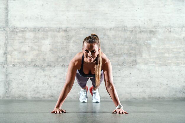 Sorridente sportiva indoeuropea positiva in abbigliamento sportivo e con coda di cavallo facendo push up al chiuso