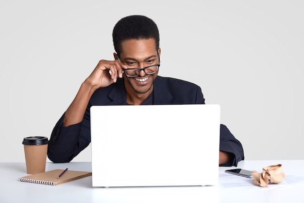 Photo smiling positive dark skinned man wears transparent glasses, focused into laptop computer, makes distant job, writes down something in notepad, drinks takeout coffee, isolated on white wall.