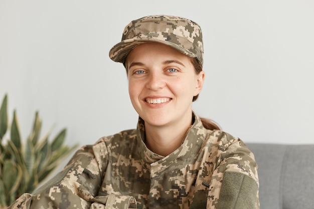 Smiling positive female soldier wearing military costume,\
posing indoor in light room, looking at camera, expressing\
optimistic emotions, being happy to come back home.