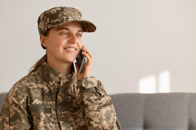 Улыбающаяся позитивная женщина-солдат в военном костюме, позирует в светлой комнате, разговаривает через смартфон, выражает оптимистические эмоции, смотрит в сторону.