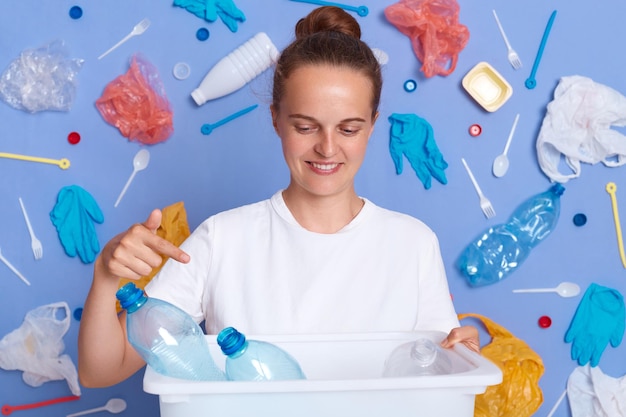 Smiling positive female with bun hairstyle holding and pointing at box of plastic bottles sorting rubbish saving our planet demonstrating serious environmental problem and ecology pollution