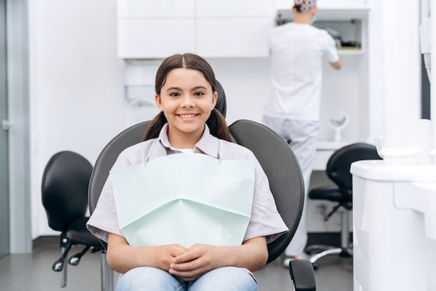 Smiling, positive girl sitting in a dental chair. On a blurred background, the dentist is preparing for work