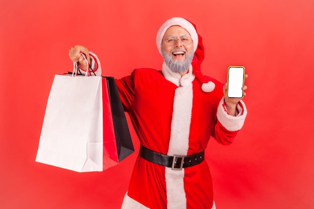 Smiling positive gray bearded santa claus holding and showing paper bags and empty screen smartphone shopping online Indoor studio shot isolated on red background