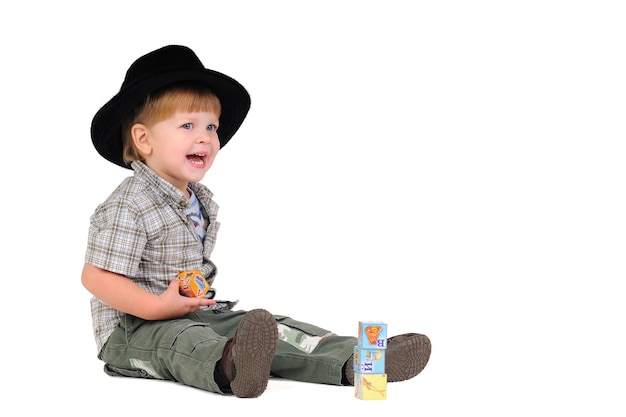 黒い帽子をかぶった笑顔のポジティブな男の子は、白い背景の床に座っている間、キューブを再生します