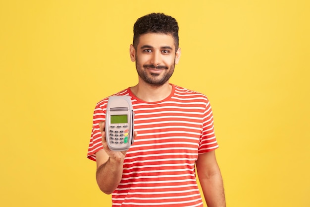 Smiling positive man with beard in striped t-shirt holding in hand pos contactless payment terminal, using paypass for purchases payment. Indoor studio shot isolated on yellow background