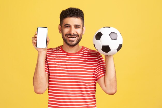 Smiling positive man with beard in striped t-shirt holding and showing white display smartphone and football ball, betting on soccer. Indoor studio shot isolated on yellow background