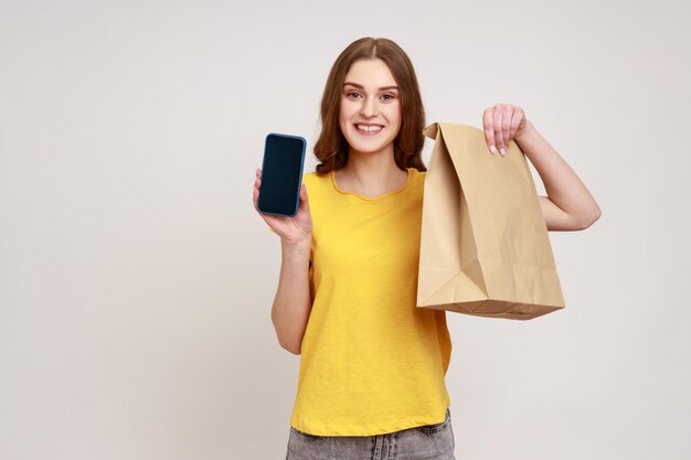 Smiling positive teenager girl in casual yellow Tshirt holding blank display smartphone and eco paper bag online shopping fast delivery marketplace Indoor studio shot isolated on gray background