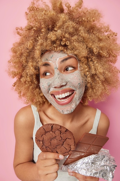 Smiling positive woman with curly bushy hair looks away
gladfully eats delicious sweet chocolate and cookie applies clay
nourishing mask on face to reduce fine lines isolated on pink
background