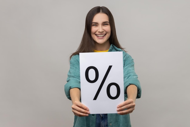 Smiling positive woman with dark hair holding paper and showing with percent sign inscription looking at camera wearing casual style jacket Indoor studio shot isolated on gray background