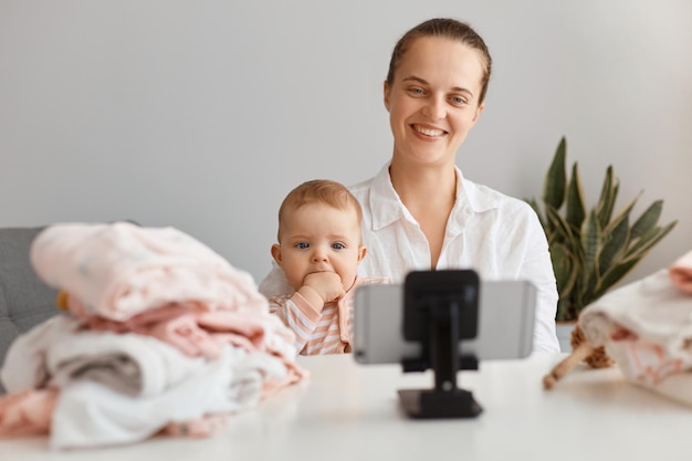 Smiling positive young female wearing white shirt broadcasting livestream at home with her charming toddler daughter, filming content for her blog, looking at phone camera.