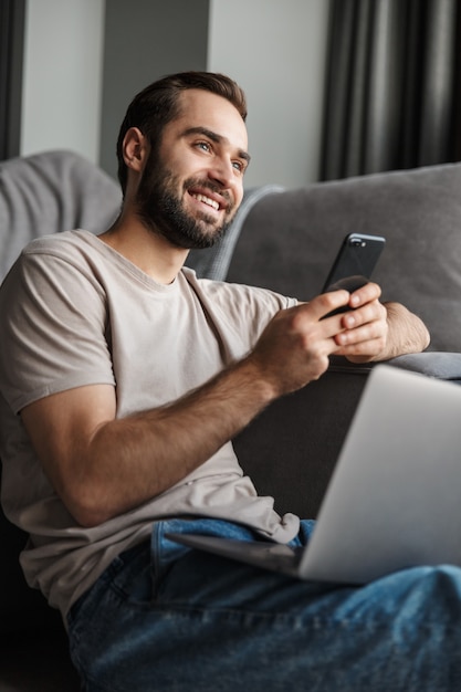 a smiling positive young man indoors at home on sofa using laptop computer chatting by mobile phone.