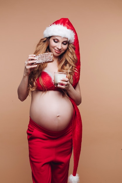 Smiling pregnant girl in Santa Claus suit eating gingerbread cookies with milk having fun