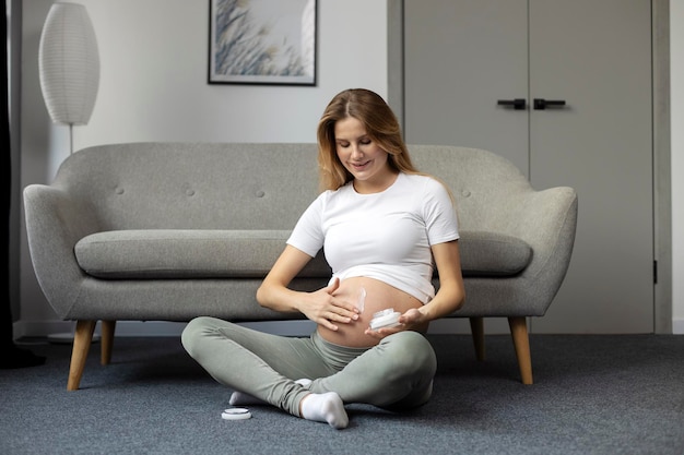 Smiling pregnant woman applying stretch mark cream on belly sitting at home Pregnancy skin care