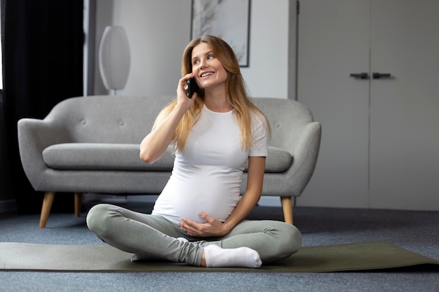 Smiling pregnant woman holding belly talking on mobile phone sitting in lotus pose at home