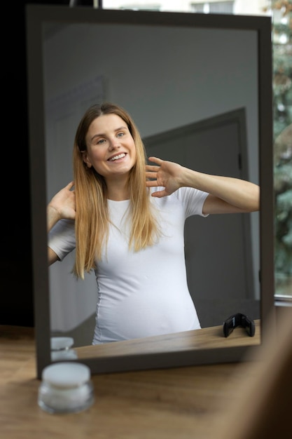 バスルームで鏡を見て笑顔の妊娠中の女性。髪の世話をする美しい女性