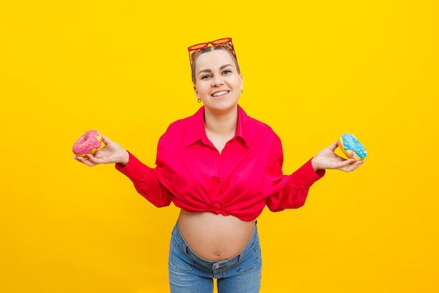 A smiling pregnant woman in a pink shirt holds donuts in her hands on a yellow background sweet food during pregnancy harmful food during pregnancy