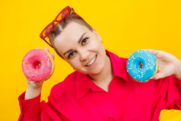 분홍색 셔츠를 입은 웃는 임산부는 노란색 배경에 도넛을 손에 들고 임신 중 단 음식 임신 중 해로운 음식
