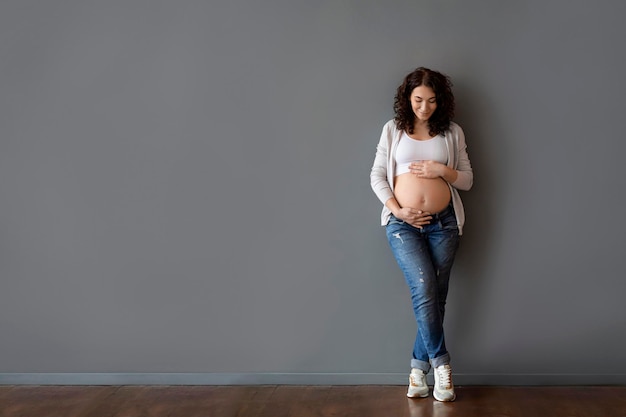 Smiling pregnant woman resting hands on belly while standing near grey wall