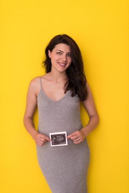 smiling pregnant woman showing ultrasound picture of her unborn baby isolated on yellow background