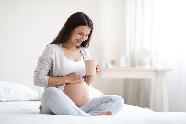 Smiling pregnant woman sitting on bed drinking tea