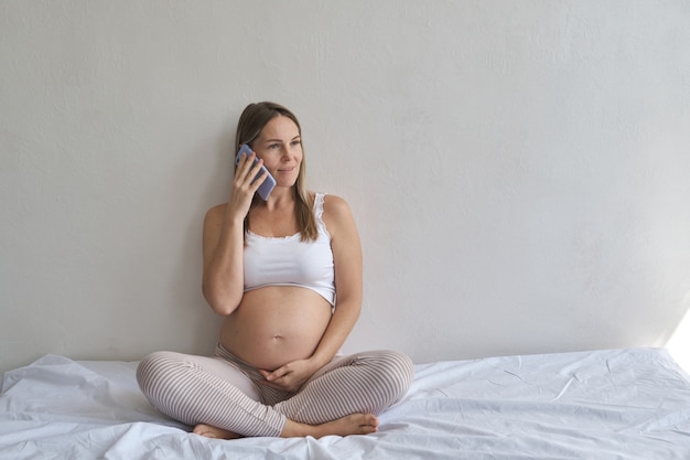 Sorridente donna incinta seduta sul letto e utilizza lo smartphone in casa