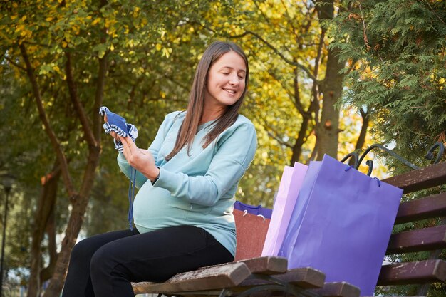 Smiling pregnant woman sitting on street bench and looking curiously at new clothes she has bought for unborn baby Mother holding baby hat in hands and looking at packages with interest