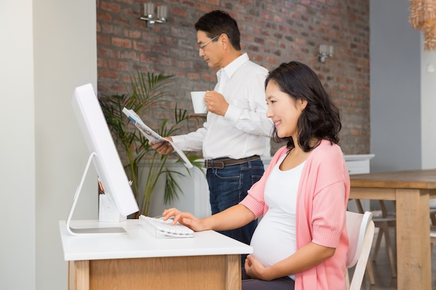 Donna incinta sorridente che per mezzo del computer portatile a casa mentre il suo marito sta leggendo il giornale