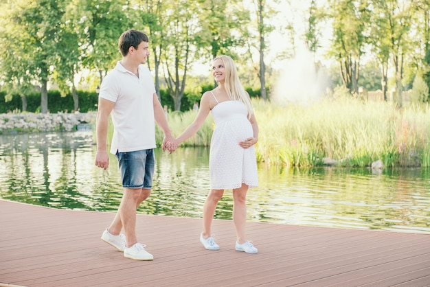 Smiling pregnant woman walking with husband on quay near the water in the evening. Free time on the nature.