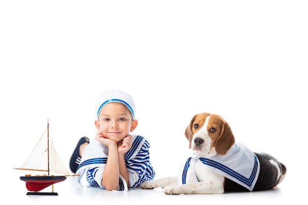 Smiling preschooler child in sailor suit with toy ship and beagle dog on white