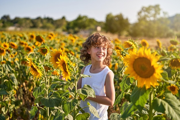 Улыбающийся ребенок-подросток смотрит в сторону и стоит в поле с цветущими подсолнухами в летний день в сельской местности