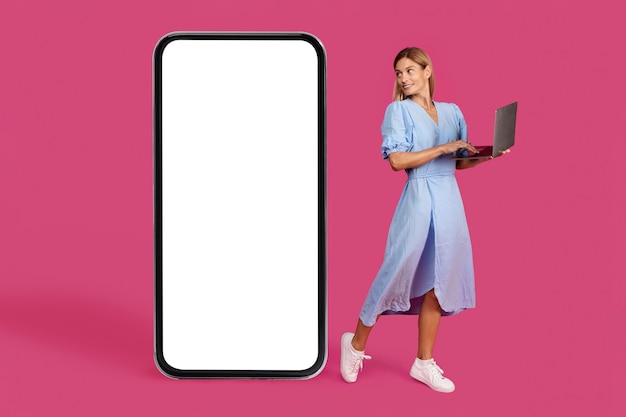 Smiling pretty adult caucasian blonde woman in dress with laptop looks at huge smartphone with blank screen