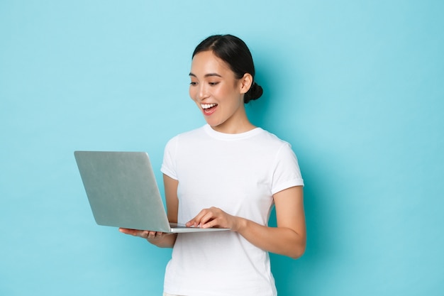 Sorridente bella ragazza asiatica in maglietta bianca, lavorando al progetto, guardando soddisfatto e felice allo schermo del laptop, freelance o internet, shopping online su un muro azzurro