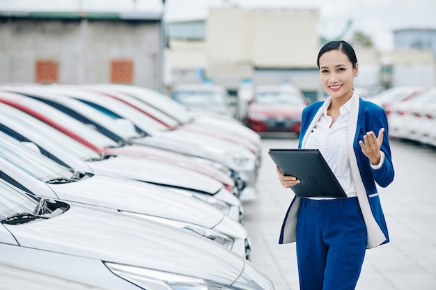 Smiling pretty Asian saleswoman inviting you to choose new car in car dealership