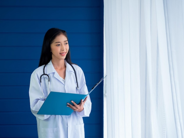 Smiling pretty asian woman doctor portrait standing on blue\
wood and white curtain background in hospital room confident asian\
young female practitioner with stethoscope holding document\
folder