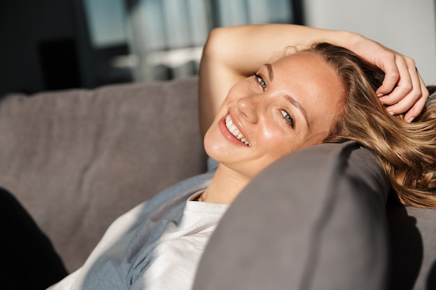 Smiling pretty blonde young woman wearing casual clothes relaxing on a couch at home