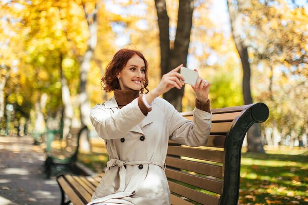 Smiling pretty caucasian millennial redhaired female in raincoat sits on bench and takes photo enjoy autumn