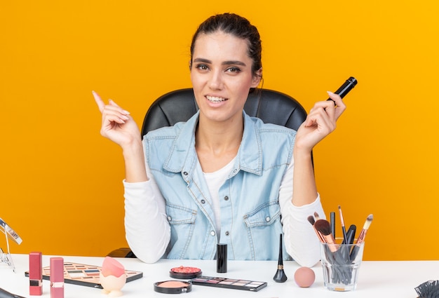 Smiling pretty caucasian woman sitting at table with makeup tools holding eyeliner and pointing at side 