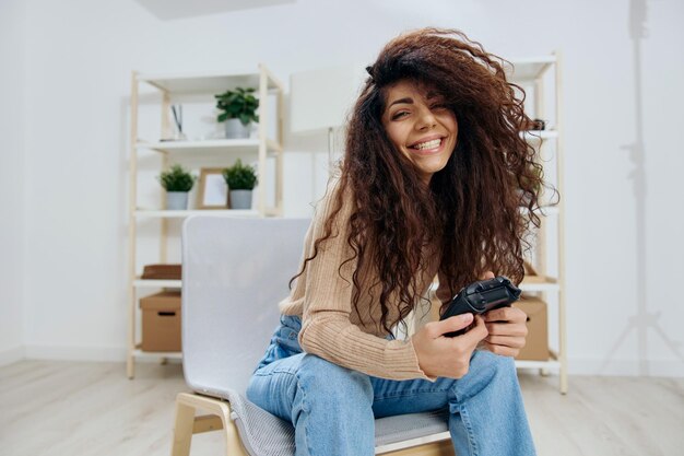 Smiling pretty enjoyed tanned curly latin winner lady hold with\
joystick gamepad in both hands sit on chair in home modern interior\
look at camera copy space mockup banner concept game platform