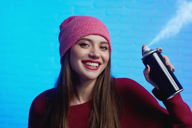 Foto femmina graziosa sorridente con capelli lunghi che portano cappello rosso e maglione che posano con lo spruzzo di pittura in sue mani