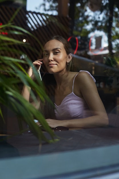 Smiling pretty lady calling by phone shot through window glass outside of cafe