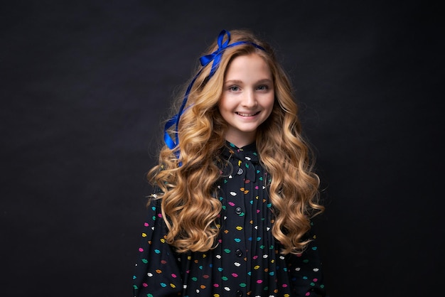 웃는 예쁜 백인 소녀 1210 검은 배경 어린이 스튜디오 초상화에 포즈를 취하는 그녀의 머리에 파란색 리본이 달린 아름다운 긴 물결 모양의 머리 어린 시절 라이프 스타일 개념