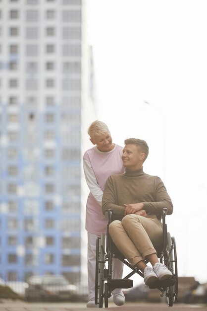 Smiling pretty mature woman with blond hair pushing wheelchair with crippled man while they walking