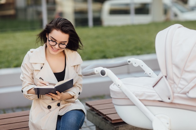 Smiling pretty mother writing something at book on bench near baby stroller in park