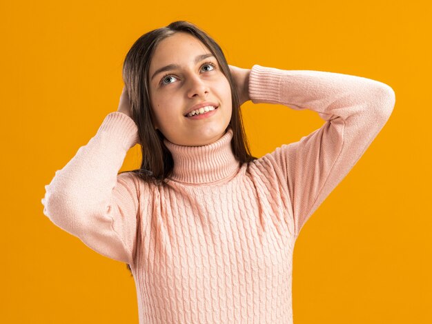 Smiling pretty teenage girl looking up keeping hands behind head isolated on orange wall