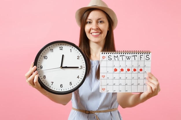 Smiling pretty woman in blue dress, hat holding round clock, periods calendar for checking menstruation days isolated on trending pink background. Medical healthcare, gynecological concept. Copy space