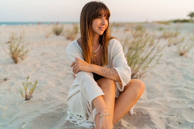 Sorridente bella donna in abito estivo bohémien con collana elegante seduta sulla sabbia