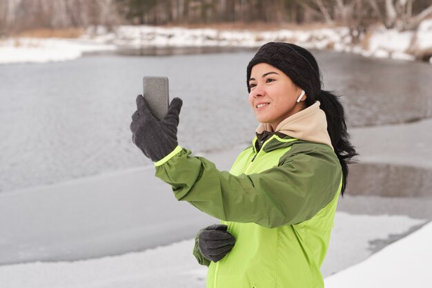 Sorridente bella donna in auricolari utilizzando smartpnone durante l'assunzione di selfie contro il lago invernale