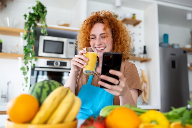 Photo smiling pretty woman looking at mobile phone and holding glass of orange juice while cooking fresh vegetables in kitchen interior at home